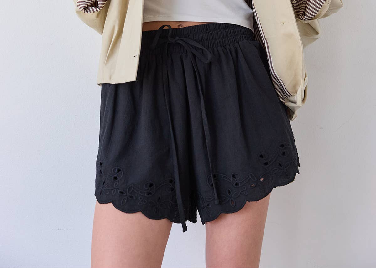 Woman wearing a beige jacket, white top, and black shorts against a plain background
