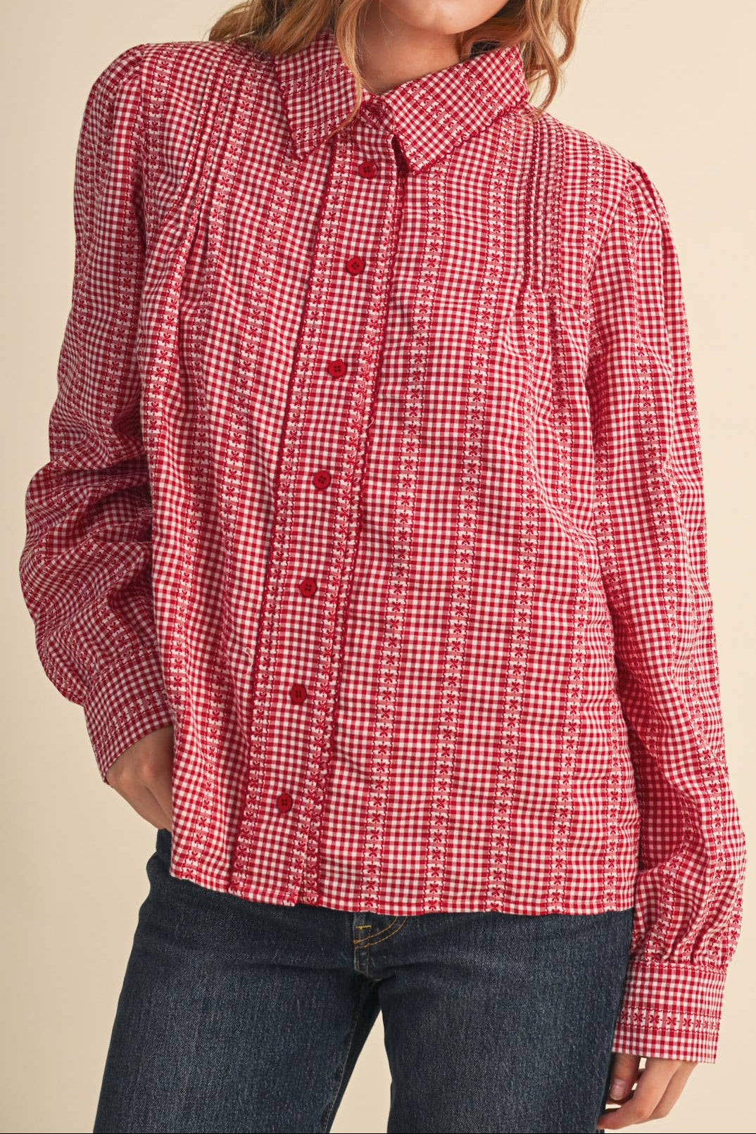 A woman wearing a red and white gingham long-sleeve button-up shirt with dark blue jeans, photographed against a solid beige background.