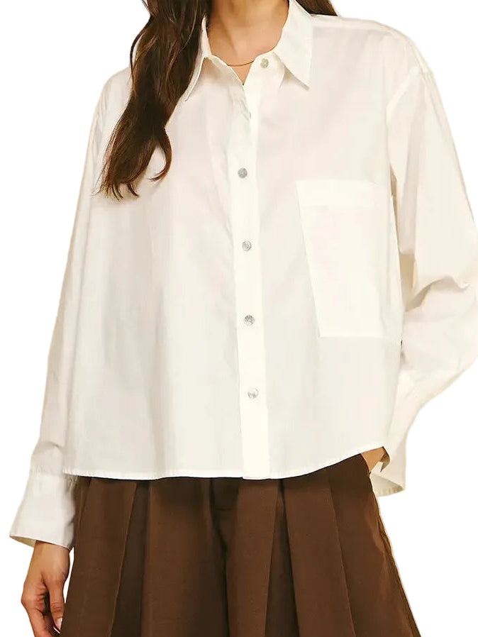 A woman modeling a white long-sleeve button-up shirt with a chest pocket, paired with brown pleated trousers, against a solid beige background.