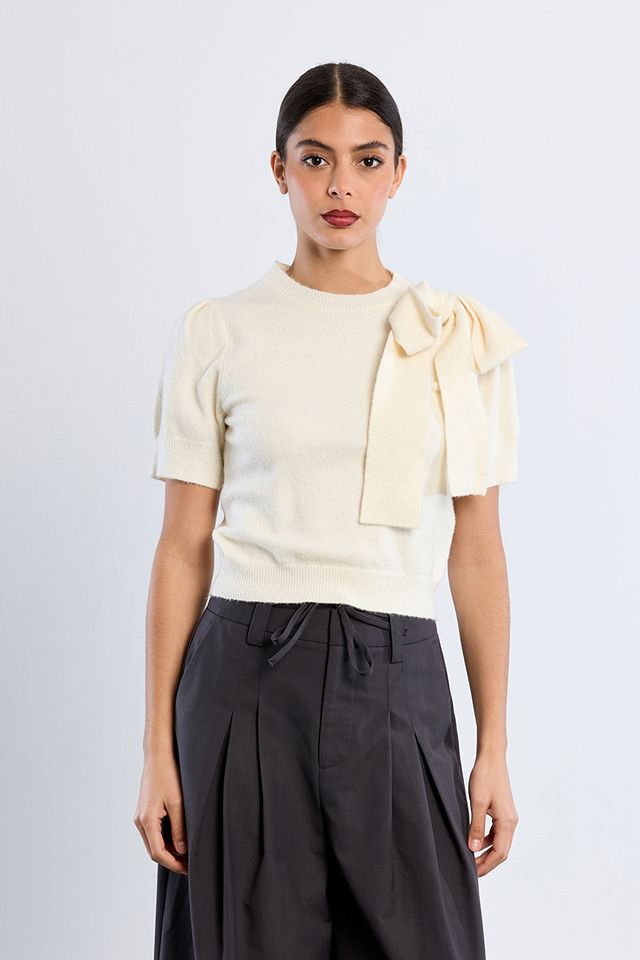 A woman modeling a cream short-sleeve knit top with a large bow detail on the shoulder, paired with dark charcoal pleated trousers, against a light gray studio background.