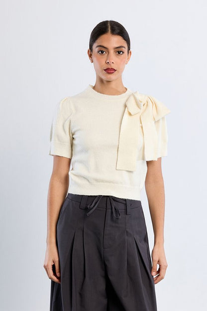 A woman modeling a cream short-sleeve knit top with a large bow detail on the shoulder, paired with dark charcoal pleated trousers, against a light gray studio background.