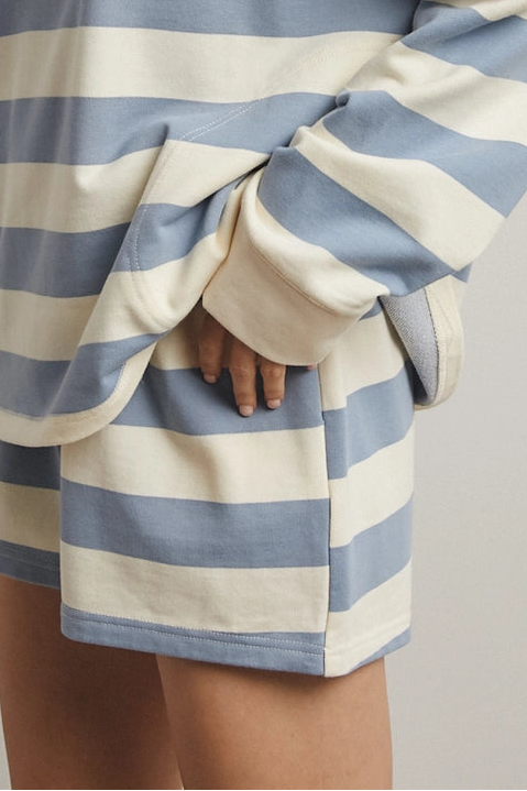 A woman wearing an oversized long-sleeve turtleneck sweatshirt with horizontal light blue and cream stripes, posed against a neutral light gray studio background.