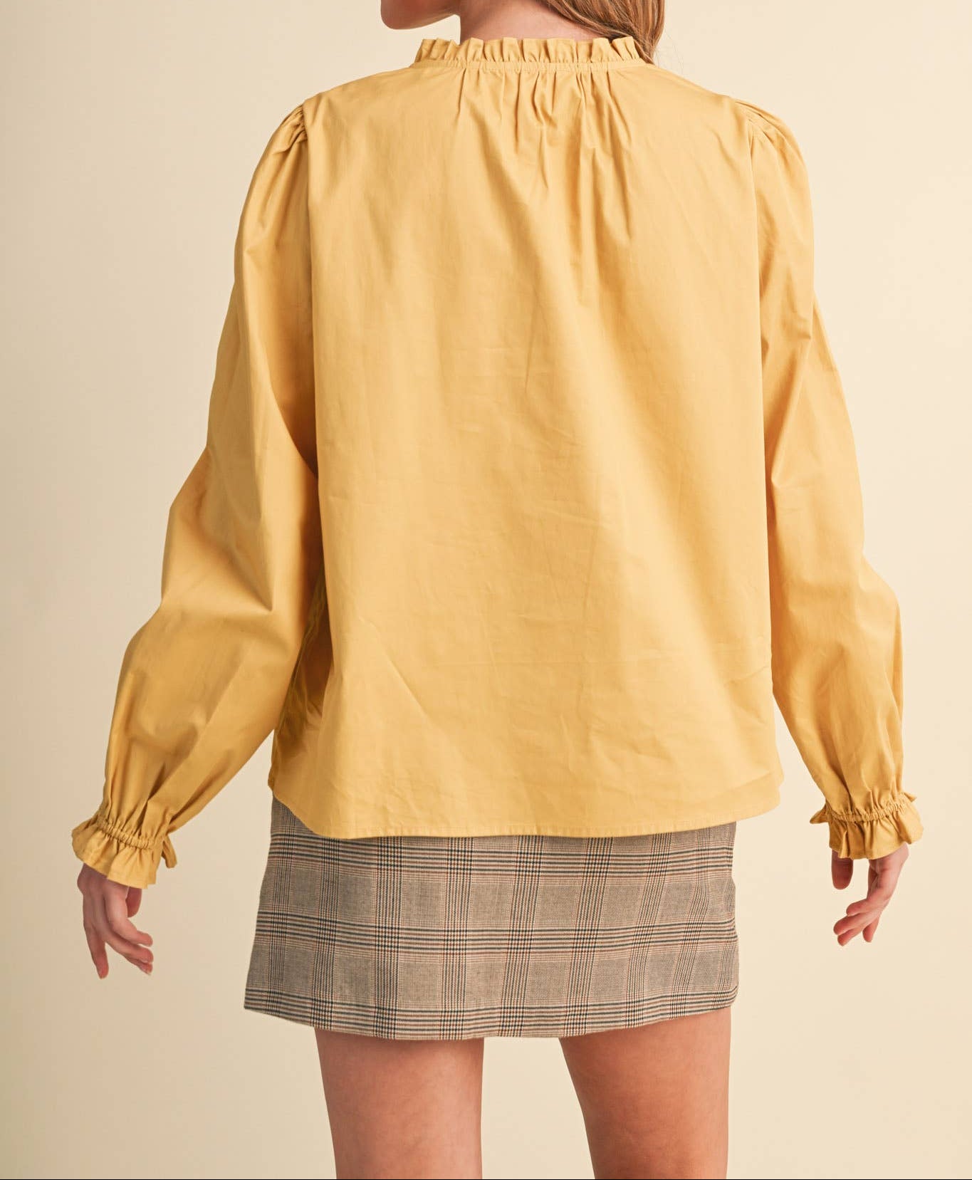 A woman shown from the back wearing a mustard yellow long-sleeve blouse with gathered ruffles at the neck and cuffs, paired with a gray plaid mini skirt, against a solid beige studio background.