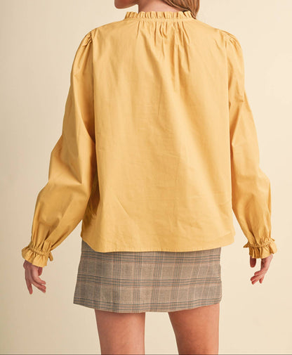 A woman shown from the back wearing a mustard yellow long-sleeve blouse with gathered ruffles at the neck and cuffs, paired with a gray plaid mini skirt, against a solid beige studio background.