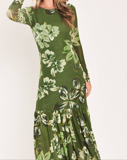 A woman wearing a long-sleeve olive green maxi dress with a large white and light green tropical floral print, posed against a plain off-white studio background.