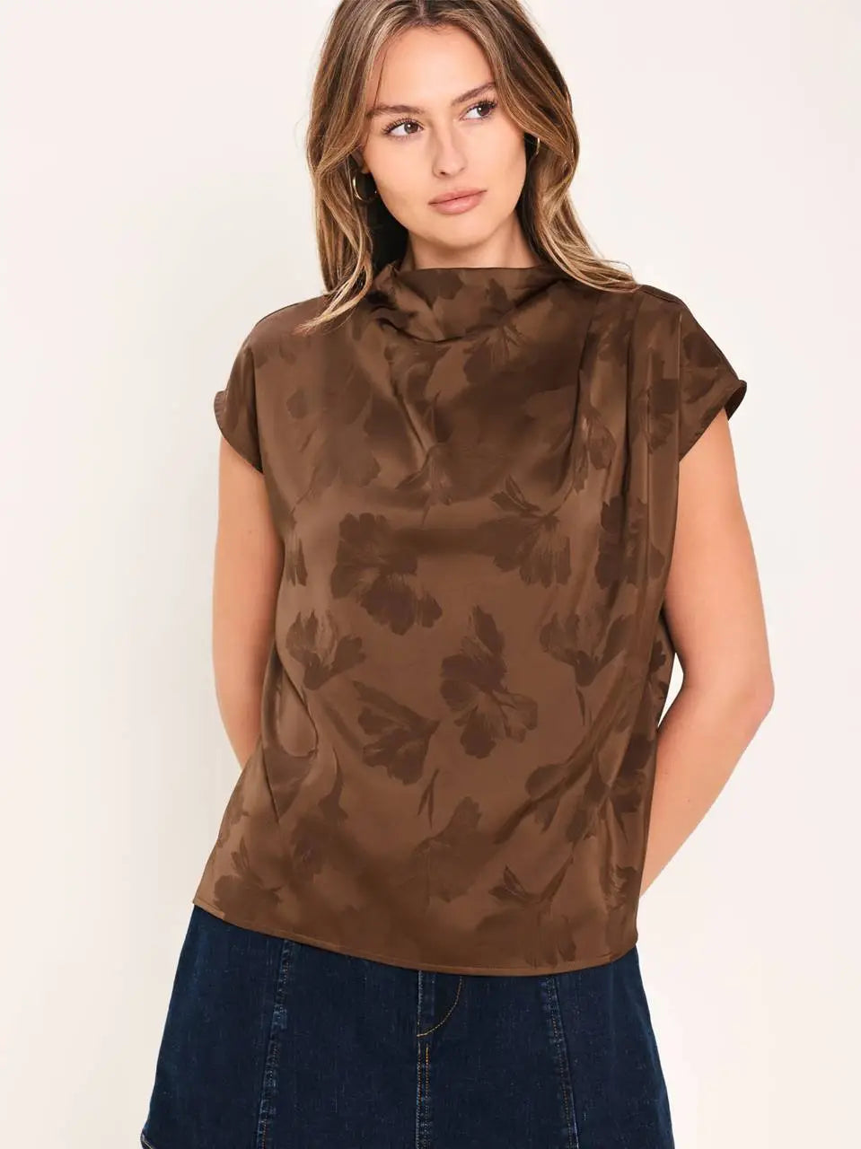 A woman wearing a brown short-sleeve top with a darker brown abstract floral pattern, paired with dark blue jeans, against a plain off-white background.