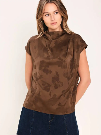 A woman wearing a brown short-sleeve top with a darker brown abstract floral pattern, paired with dark blue jeans, against a plain off-white background.