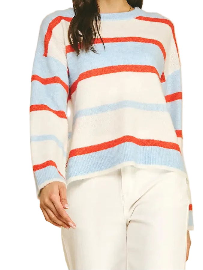 A woman modeling a long-sleeve knit sweater with horizontal stripes in light blue, coral red, and off-white, paired with off-white pants against a solid beige background.