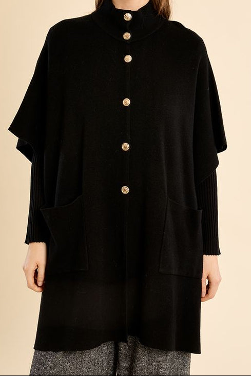 A woman modeling a black high-neck, long-sleeved knit poncho-style cardigan with gold buttons down the front, set against a solid beige background.