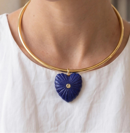 Gold necklace with a blue heart-shaped pendant worn by a person in a white shirt.