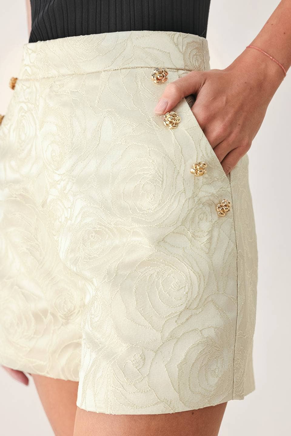 Close-up of a model wearing a cream-colored textured mini skirt with gold floral buttons and a side pocket.