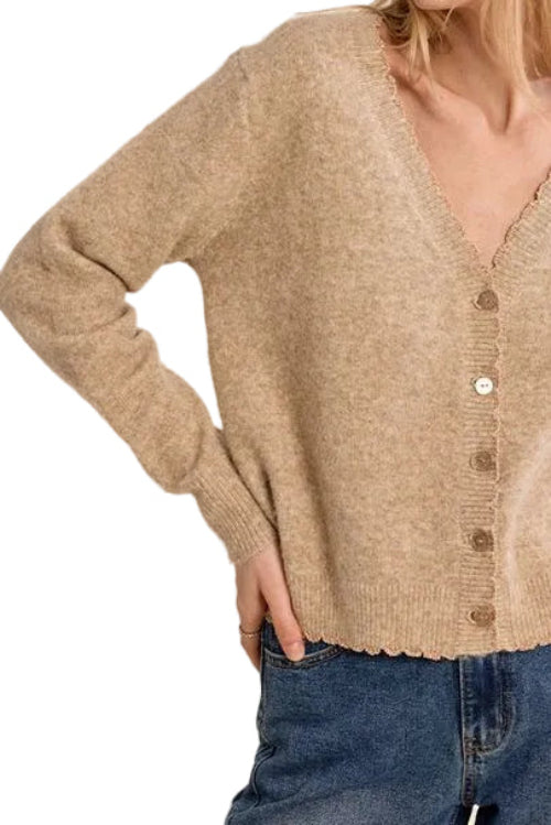 A woman wearing a beige knit cardigan sweater with a scalloped V-neck and button-front closure, paired with blue jeans, against a plain white studio background.