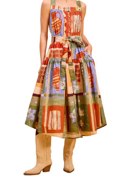 A woman models a sleeveless midi dress with a colorful patchwork print in red, green, blue, and orange, styled with beige boots against a solid warm white studio backdrop.