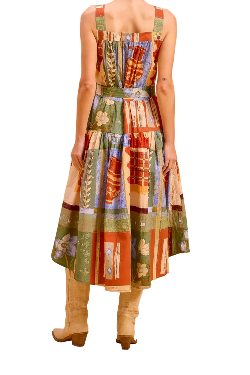 A woman stands with her back to the camera wearing a sleeveless midi dress with a colorful patchwork print in green, red, orange, and beige, paired with beige knee-high boots, set against a solid warm beige studio backdrop.