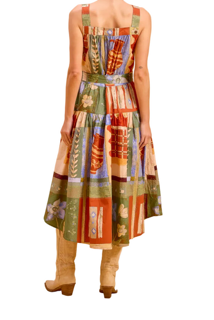 A woman stands with her back to the camera wearing a sleeveless midi dress with a colorful patchwork print in green, red, orange, and beige, paired with beige knee-high boots, set against a solid warm beige studio backdrop.