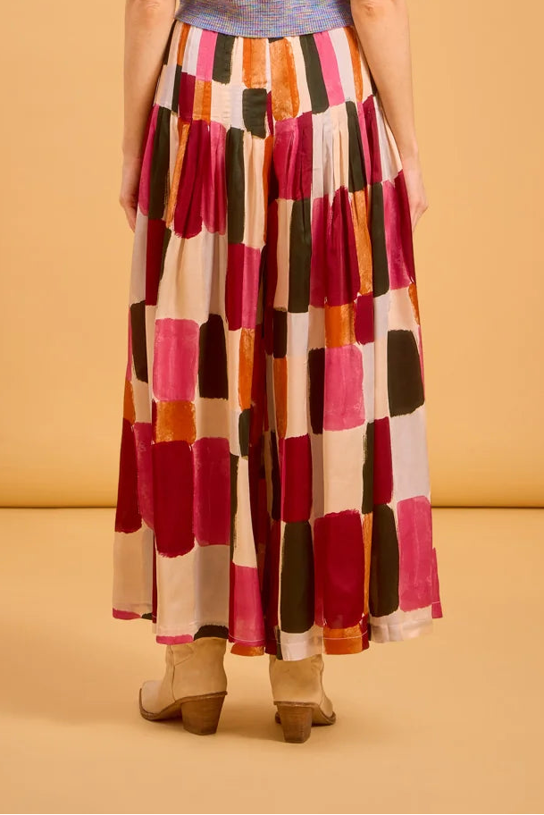 A woman viewed from behind wearing a short-sleeve knit top with a multicolor stripe pattern and a long pleated pants with a large patchwork block pattern in pink, red, orange, cream, and dark green. She is standing against a solid warm beige studio background.