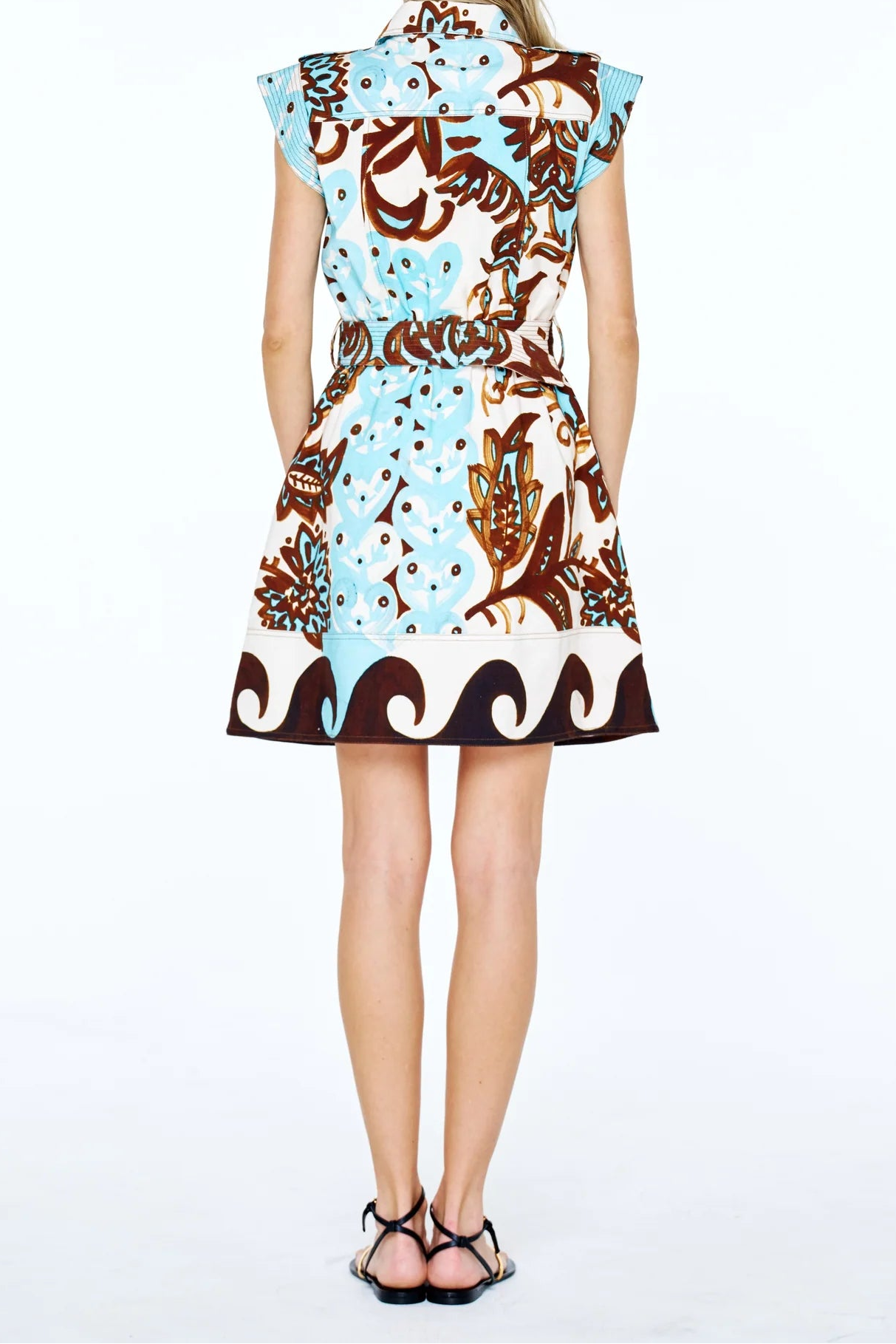 A woman viewed from behind wearing a sleeveless, knee-length dress with a brown and light blue abstract pattern on a white base, paired with black strappy sandals, against a plain white studio background.