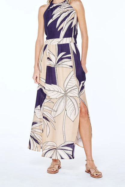 A woman modeling a sleeveless halter-neck maxi dress with a tropical palm leaf print in navy blue and beige, paired with brown strappy sandals against a white studio background.