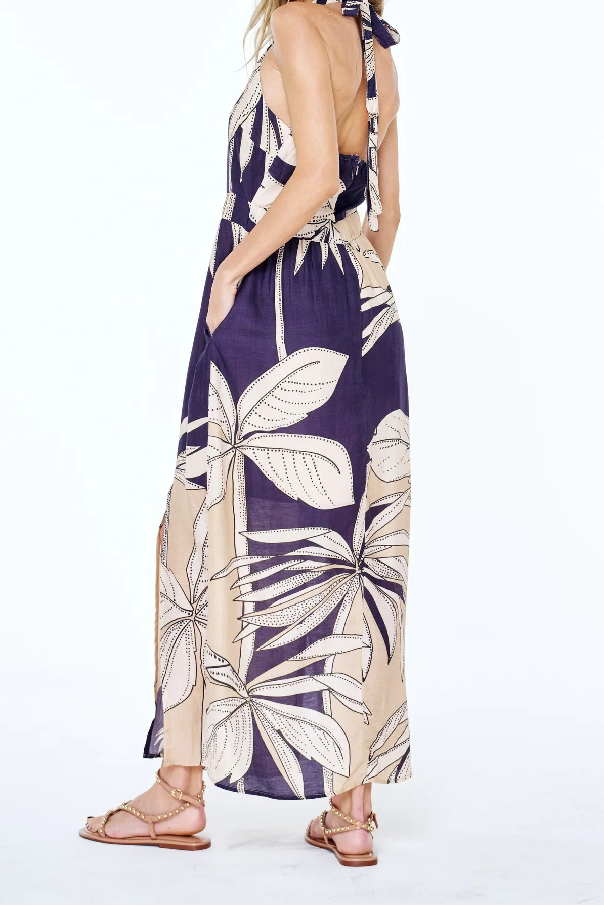 A woman modeling a sleeveless halter maxi dress with a large tropical palm leaf print in navy blue and beige, shown from a side-back angle against a plain white studio background.
