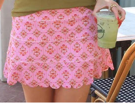 Woman in green tank top and pink skirt walking outdoors near a building.