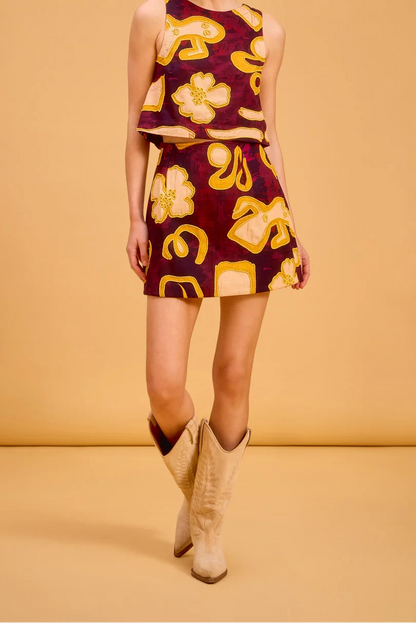 A woman modeling a sleeveless two-piece set consisting of a cropped tank top and a matching mini skirt, both in a deep burgundy fabric with a bold yellow and gold abstract floral and geometric pattern. She is wearing beige cowboy boots against a solid warm tan studio backdrop.