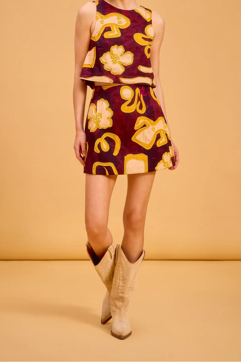 A woman modeling a sleeveless two-piece set consisting of a cropped tank top and a matching mini skirt, both in a deep burgundy fabric with a bold yellow and gold abstract floral and geometric pattern. She is wearing beige cowboy boots against a solid warm tan studio backdrop.