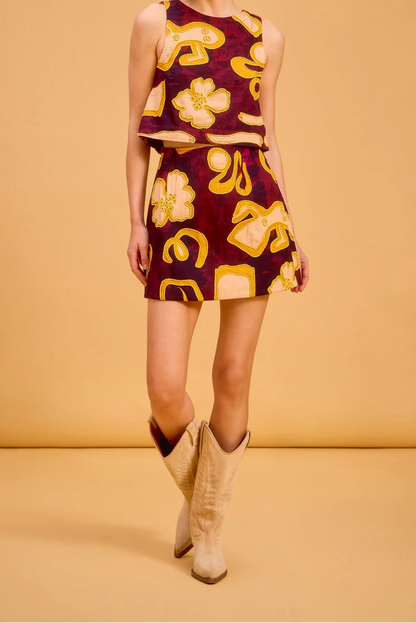 A woman modeling a sleeveless two-piece set consisting of a cropped tank top and a matching mini skirt, both in a deep burgundy fabric with a bold yellow and gold abstract floral and geometric pattern. She is wearing beige cowboy boots against a solid warm tan studio backdrop.