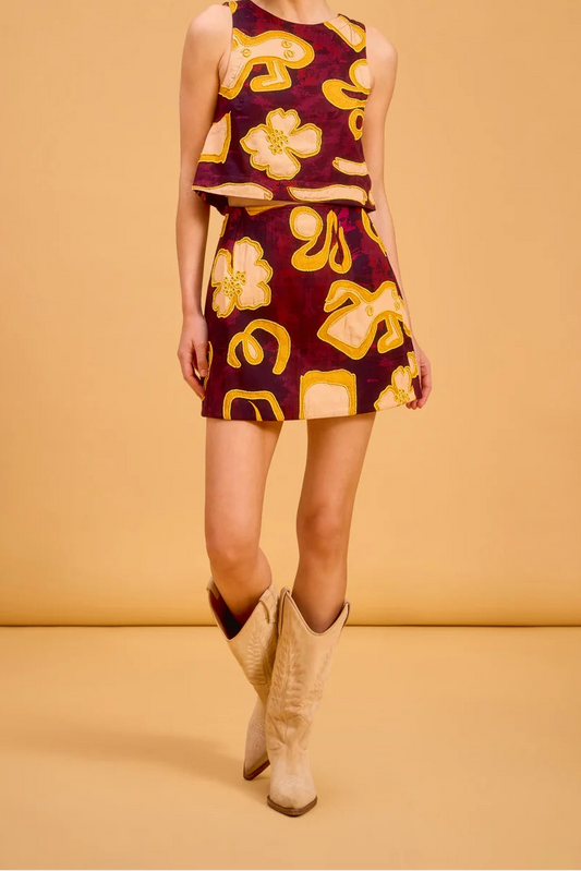 A woman modeling a sleeveless two-piece set consisting of a cropped tank top and a matching mini skirt, both in a deep burgundy fabric with a bold yellow and gold abstract floral and geometric pattern. She is wearing beige cowboy boots against a solid warm tan studio backdrop.