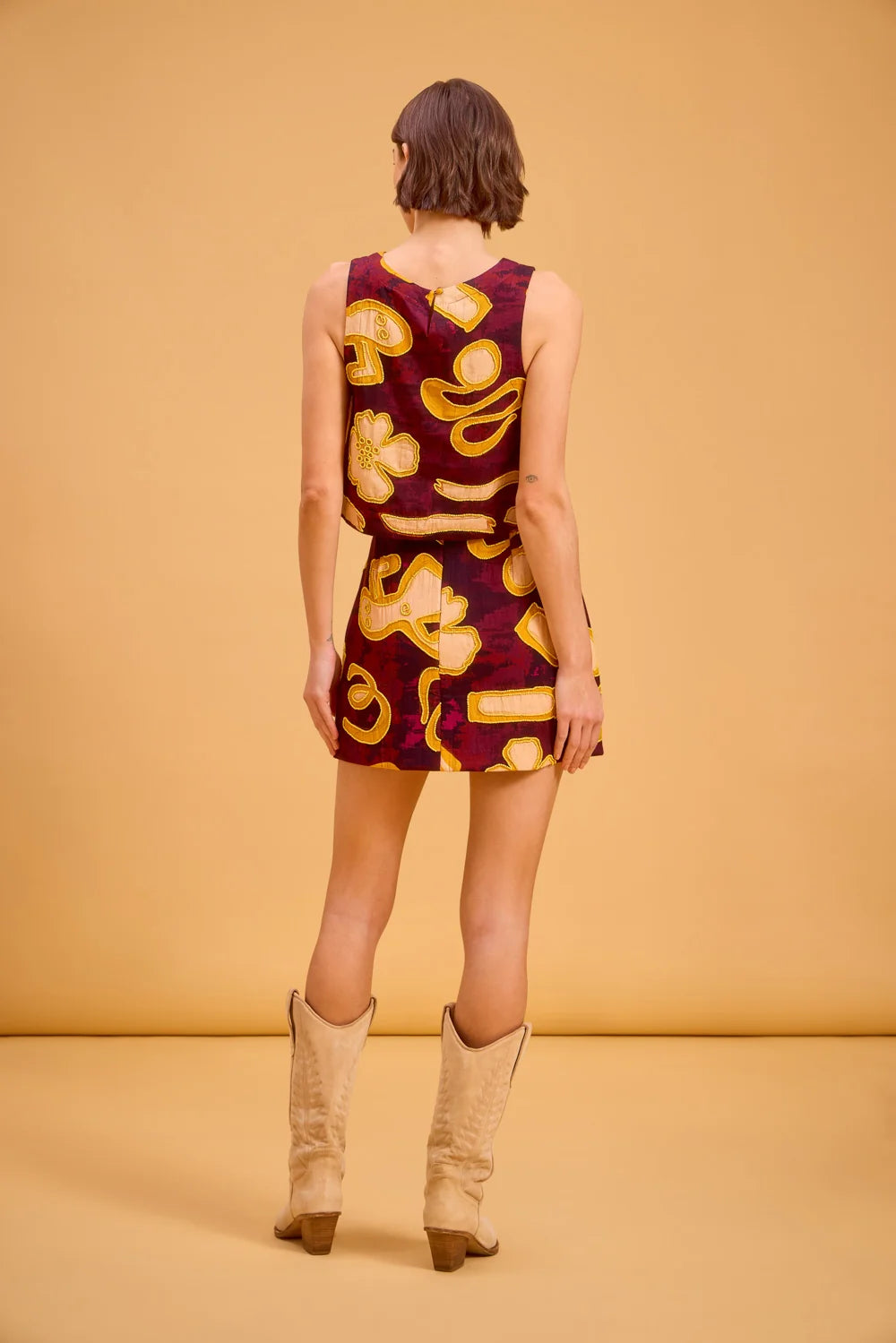 A woman stands with her back to the camera wearing a sleeveless mini dress with a retro paisley pattern in burgundy and yellow, paired with beige cowboy boots, against a solid warm beige studio backdrop.