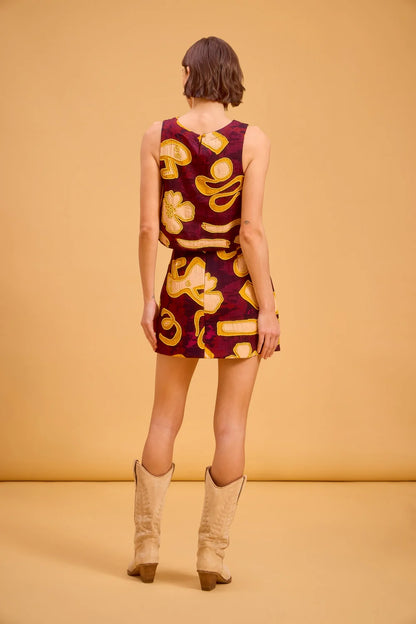A woman stands with her back to the camera wearing a sleeveless mini dress with a retro paisley pattern in burgundy and yellow, paired with beige cowboy boots, against a solid warm beige studio backdrop.