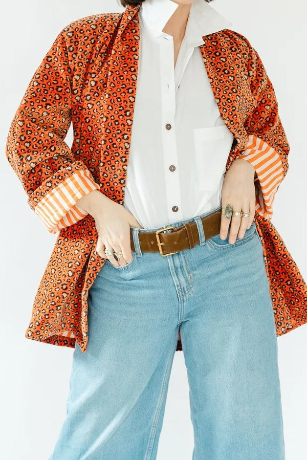 A model posing in a studio setting wearing a rust-orange patterned cardigan over a white button-down shirt, paired with light-wash wide-leg denim jeans, a brown belt, and brown suede boots.