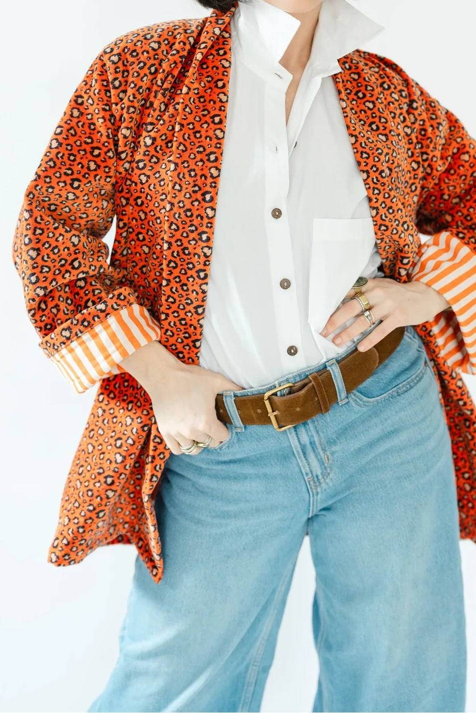 A woman posing in a retro-style outfit featuring a rust-orange patterned jacket over a white button-down shirt, paired with light blue jeans and orange-tinted sunglasses, set against a plain white background.