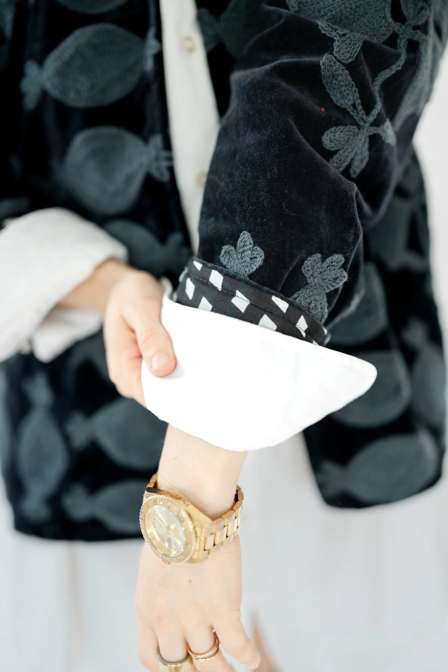 A close-up of a person wearing a black velvet jacket with a gray floral pattern, a white shirt, and a gold watch on the wrist.