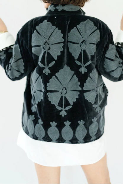 A person shown from behind wearing a black velvet jacket with a gray floral pattern, layered over a white shirt and white skirt, against a plain white background.