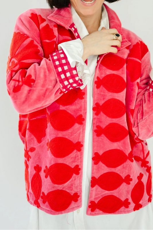 A woman posing in a bright pink fleece jacket with a red candy print, a white collared shirt, and a white skirt against a light gray studio background.