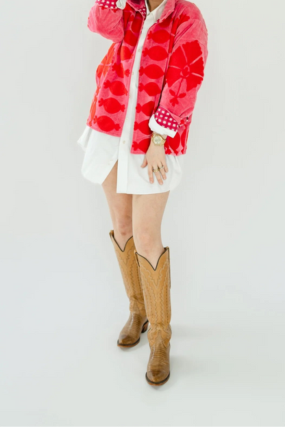 A woman modeling a vibrant pink and red patterned jacket, styled with a white shirt, white skirt, and brown cowboy boots, set against a plain white studio background.