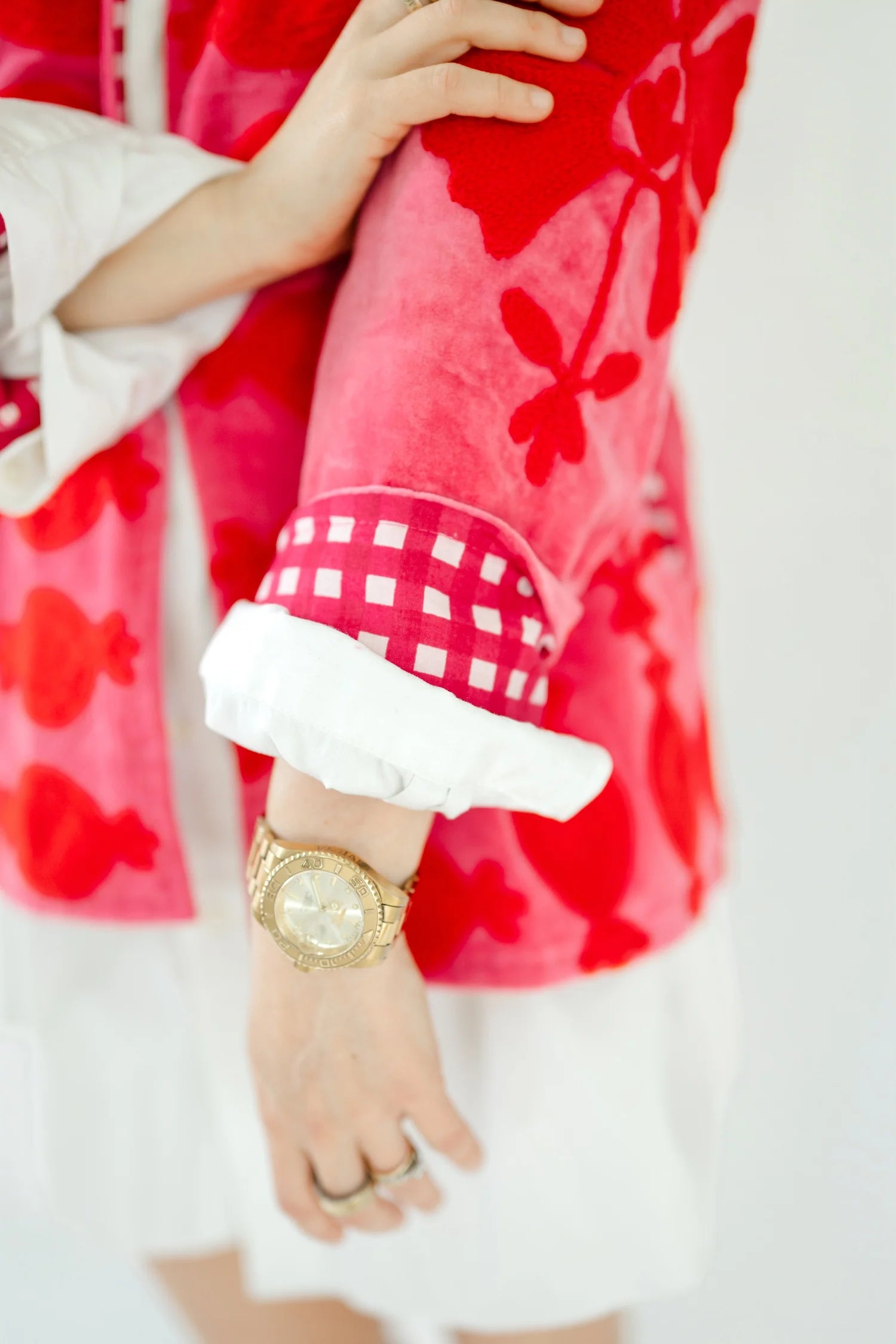 A close-up lifestyle shot of a person wearing a plush pink and red floral jacket with a white gingham cuff and white lining, paired with a white skirt and a gold watch.