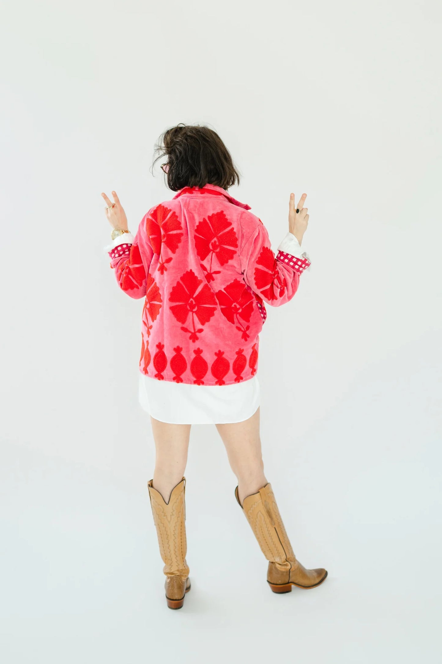 A person shown from the back wearing a bright pink and red patterned jacket, a white mini skirt, and tan cowboy boots against a plain white studio background.