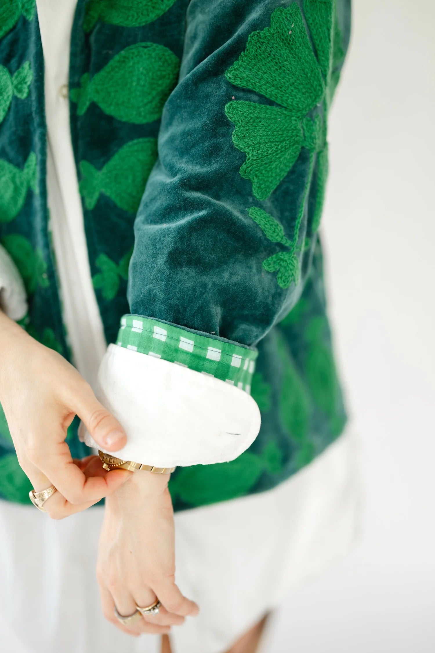 Close-up of a person wearing a green velvet jacket with a pattern, a white shirt underneath, and a green and white gingham cuff, with hands adjusting a gold watch.