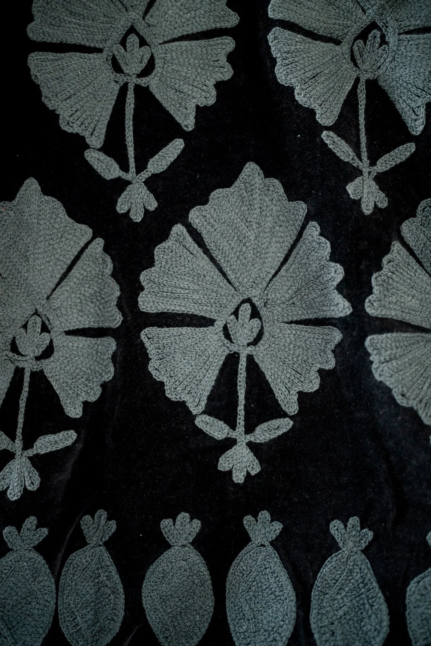 Close-up of a black fabric with a repeating light gray floral and leaf pattern, plus a row of small leaf-like motifs along the bottom edge.