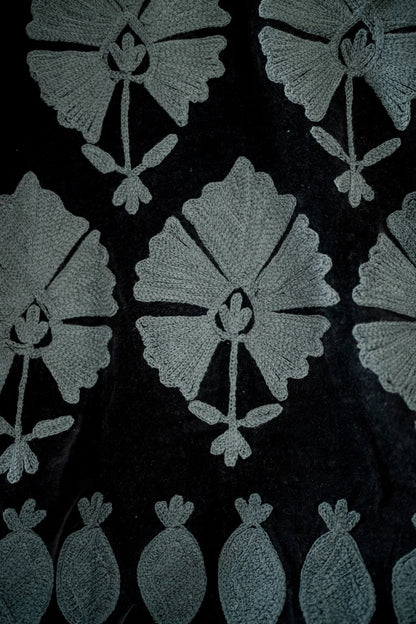 Close-up of a black fabric with a repeating light gray floral and leaf pattern, plus a row of small leaf-like motifs along the bottom edge.