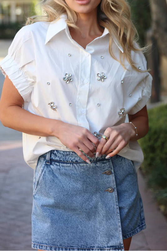 Woman wearing a white blouse with embellishments and a denim skirt outdoors.