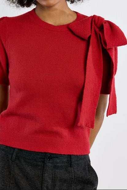 A woman with dark curly hair wearing a bright red short-sleeve knit top with a large bow detail on the right shoulder, paired with dark gray pants, against a light gray studio background.