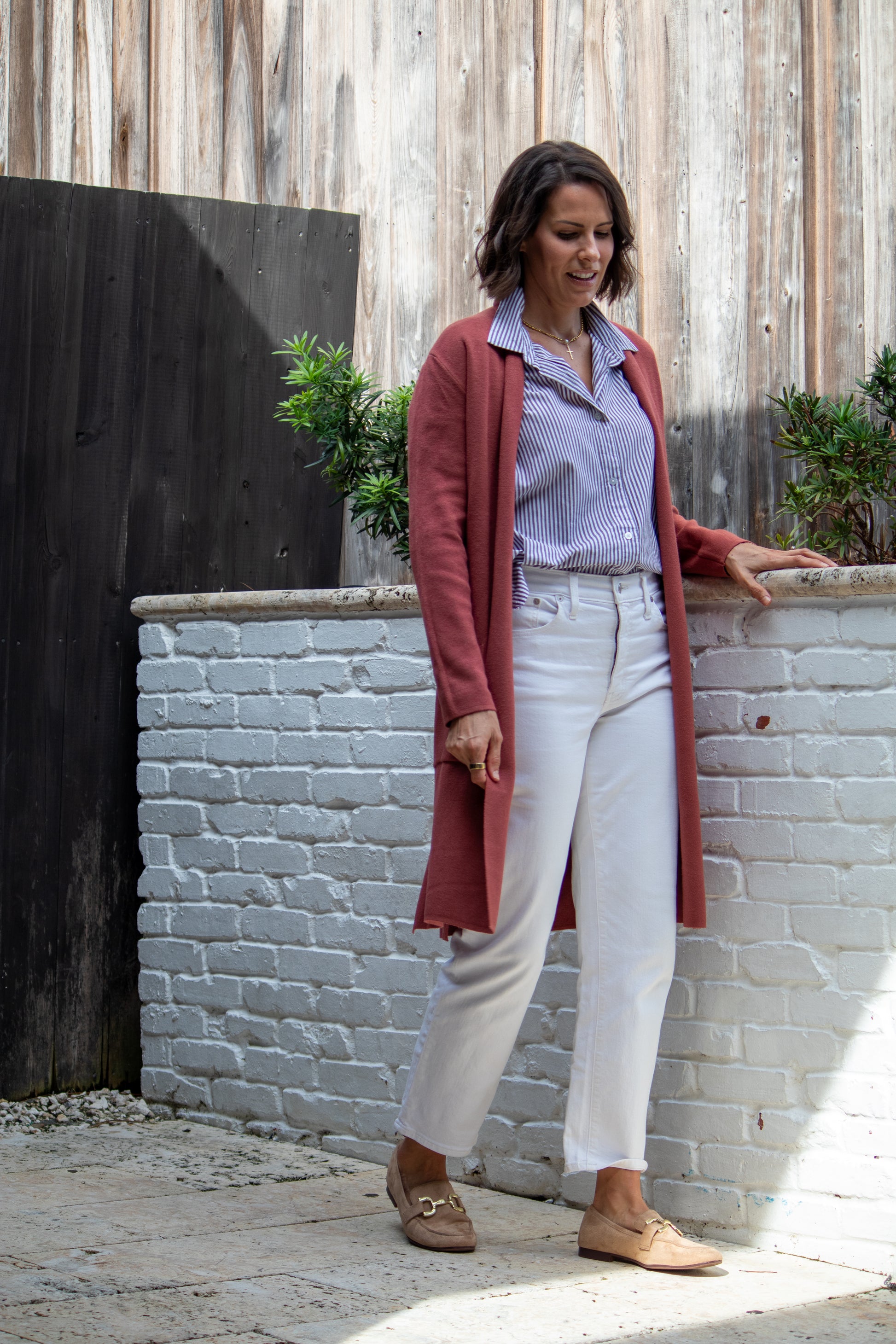 Woman standing against a white brick wall wearing a long red cardigan, light blue shirt, white pants, and brown shoes.
