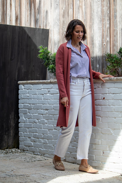 Woman standing against a white brick wall wearing a long red cardigan, light blue shirt, white pants, and brown shoes.