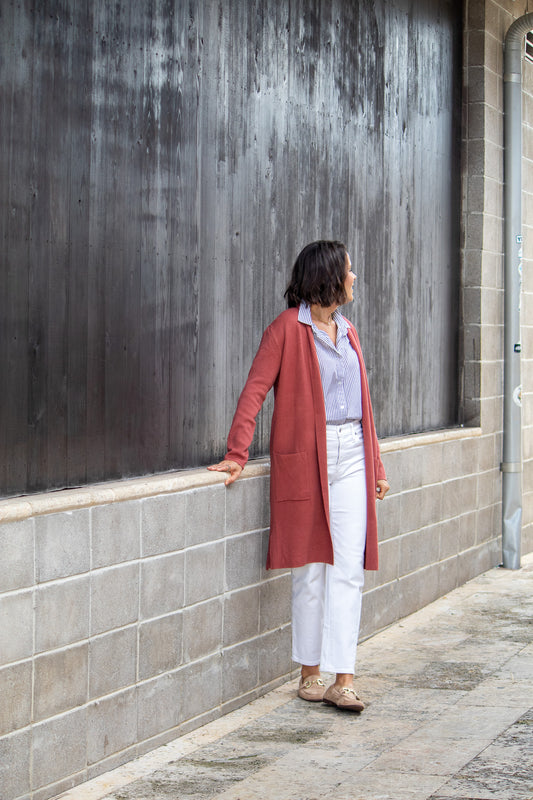 Woman standing against a textured wall wearing a red coat and white pants.