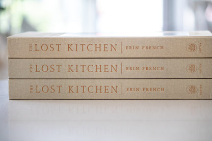 Three copies of 'The Lost Kitchen' by Erin French stacked on a light surface with a blurred background.