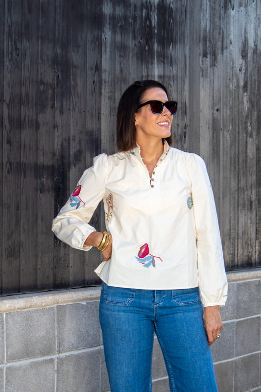 Woman wearing a white blouse with floral embroidery and blue jeans against a wooden wall.