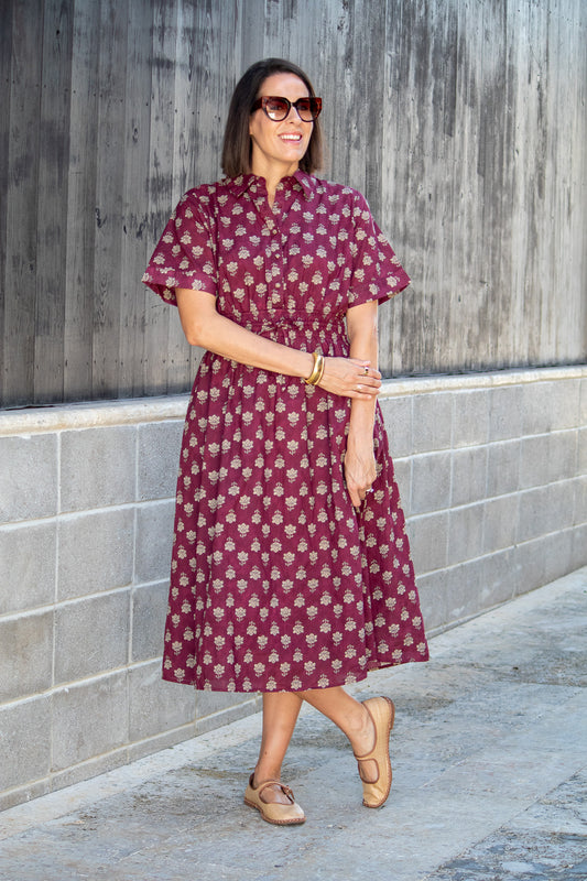 Woman wearing a maroon dress with white floral pattern standing against a gray wall.