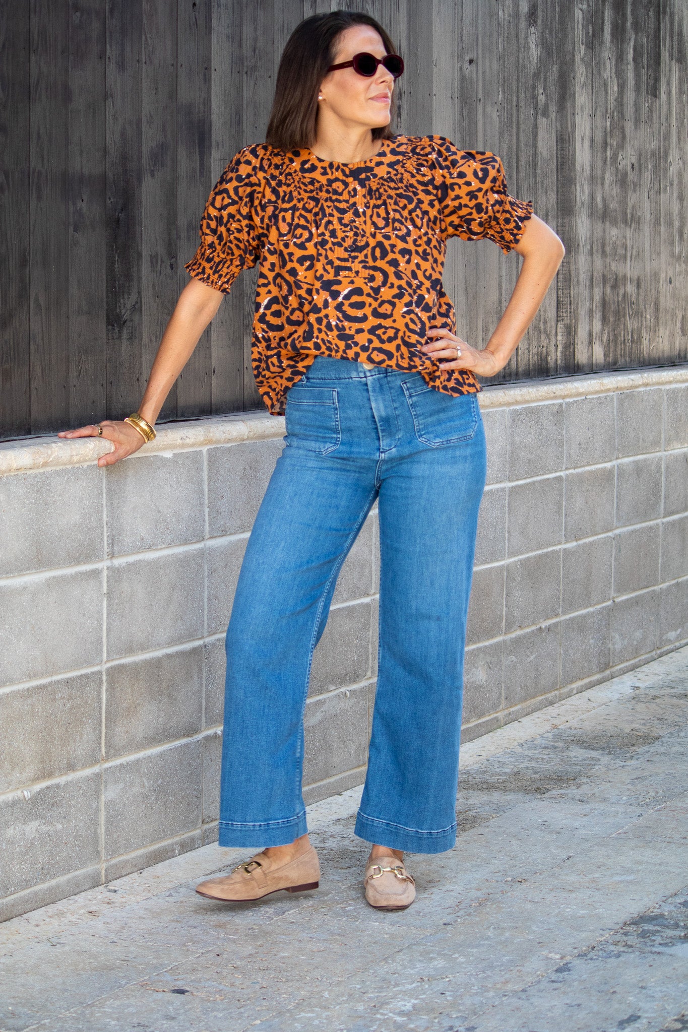Woman wearing a leopard print top and blue jeans standing against a gray wall.